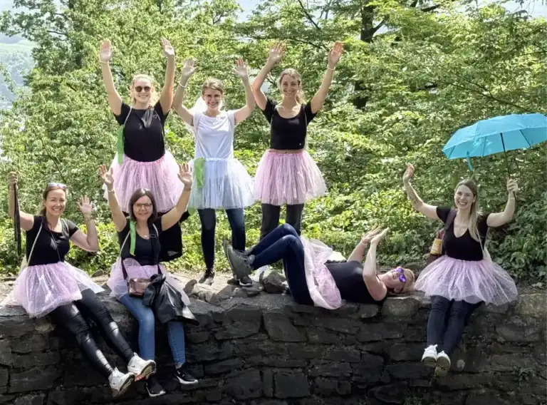 JGA-Event-Schnitzeljagd-Escape-Game-Outdoor Gruppe aus Frauen beim Junggesellenabschied auf einer Schnitzeljagd mit den Händen in der Luft auf einer Mauer