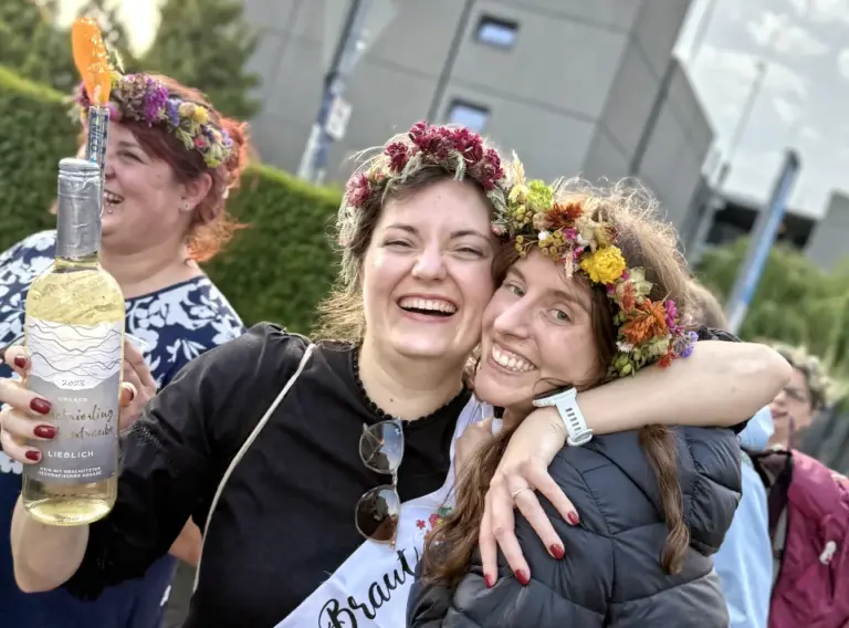 JGA-Junggesellenabschied-Schatzsuche-Schnitzeljagd Eine Braut mit ihrer Freundin im Arm bei der Siegerehrung auf einer Schnitzeljagd in Bonn, die beide Blumenkränze auf dem Kopf tragen.