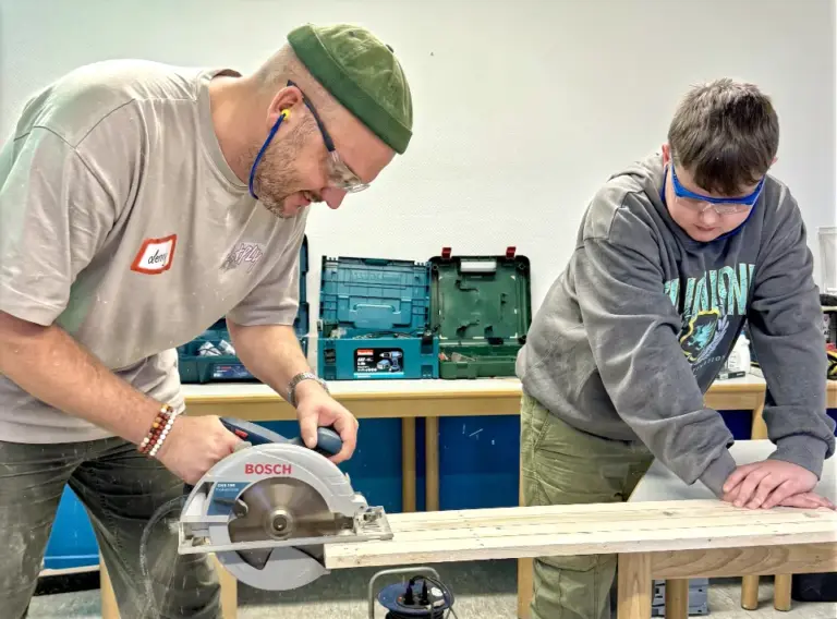 Ein Betreuer sägt mit einem Kind auf Ferienfreizeit Essen Holz für den Bau eines Holzschlittens.
