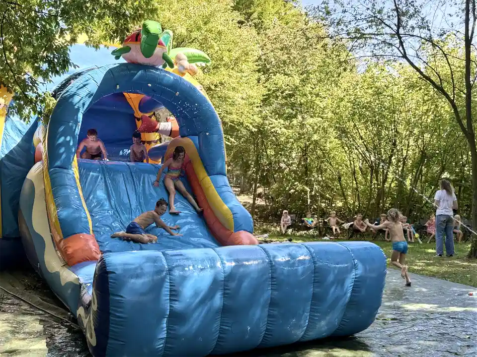 Kinder rutschen lachend eine große blaue aufblasbare Wasserrutsche hinunter, während andere im Hintergrund im Schatten sitzen und spielen – Sommerferienbetreuung in der DJH Jugendherberge Essen.