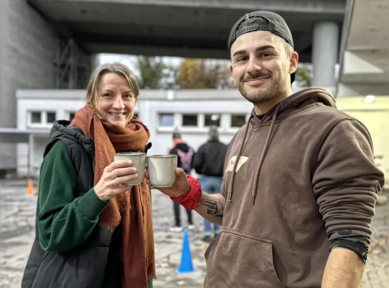 Glühweinevent Glühweinwanderung Glühweinrallye Zwei Personen stoßen bei einer Outdoor-Weihnachtsfeier mit warmen Getränken in Metallbechern an. Beide stehen lächelnd im Freien vor einer winterlichen Event-Kulisse mit Parcours-Elementen und bunten Markierungshütchen. Die Szene vermittelt eine warme, freundliche Stimmung während eines winterlichen Teamevents