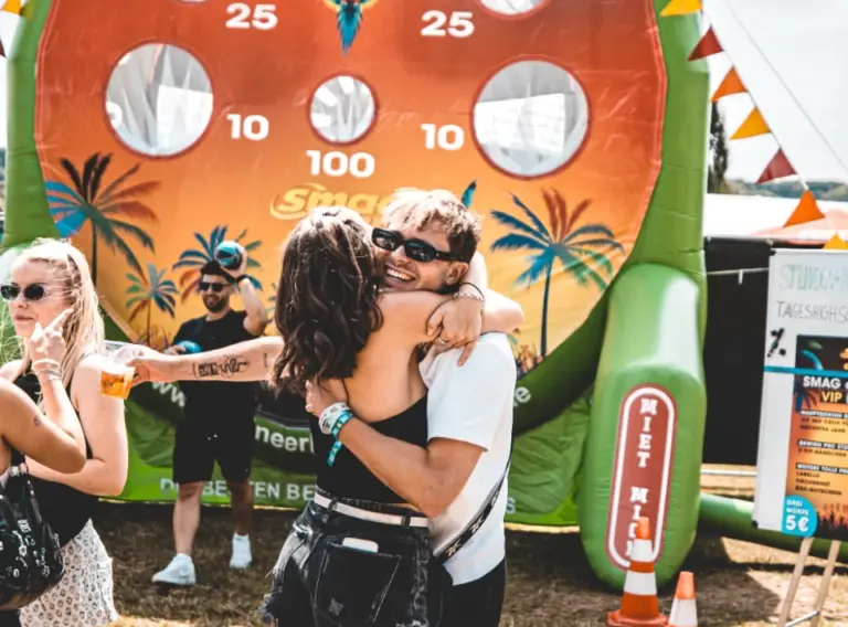 Lochwand-Fußballdarts-5.0-Meter-SMAG-Sundance-Festival-2025 Aufgeblasenes Fußball-Darts mit einer riesigen Lochwand auf dem SMAG Sundance Festival 2025 am Seaside Beach Baldeney, bei dem sich im Vordergrund zwei Teilnehmer in den Armen halten.