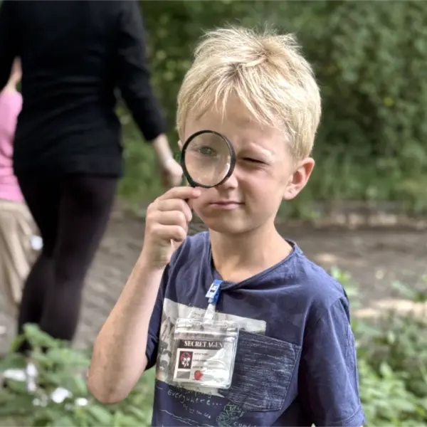Junge auf einem Kindergeburtstag bei einer Schatzsuche, wobei dieser mit einer Lupe in der Hand ausgestattet ist.
