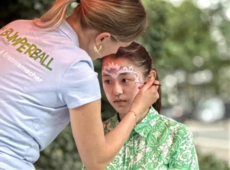 Sommerfest-Jahrmarkt-Teambuilding-Event-Firmenfeier-Kinderschminken Eine Mitarbeiter aus dem Team von Deine Erlebnismacher schminkt eine Kind beim Kinderschminken auf einer Firmenfeier in Hilden.