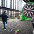 Teilnehmende einer Outdoor-Weihnachtsfeier spielen eine Fußball-Darts-Challenge im Rahmen einer Winter-Team-Olympiade. Eine große aufblasbare Dartscheibe steht als Zielwand bereit, während ein Spieler einen Ball in Richtung Ziel kickt. Bunte Wimpelketten, Markierungshütchen und winterliche Outdoor-Atmosphäre sorgen für ein lebhaftes Teamevent
