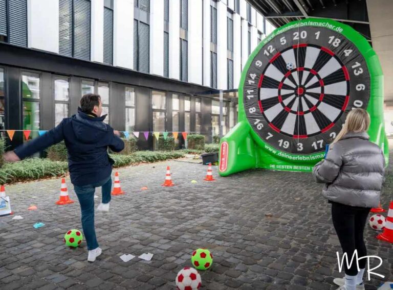 Weihnachtsfeier Outdoor-Winter-Team-Olympiade Teilnehmende einer Outdoor-Weihnachtsfeier spielen eine Fußball-Darts-Challenge im Rahmen einer Winter-Team-Olympiade. Eine große aufblasbare Dartscheibe steht als Zielwand bereit, während ein Spieler einen Ball in Richtung Ziel kickt. Bunte Wimpelketten, Markierungshütchen und winterliche Outdoor-Atmosphäre sorgen für ein lebhaftes Teamevent