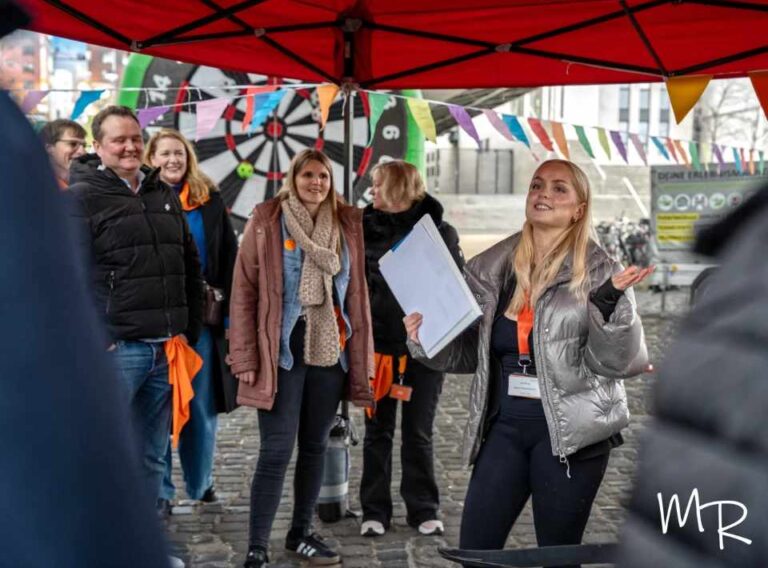 Weihnachtsfeier Outdoor-Winter-Team-Olympiade-Winterliche Teambuilding Event Eine Gruppe von Teilnehmenden steht unter einem roten Pavillon bei einer Outdoor-Weihnachtsfeier. Eine Spielleiterin erklärt lächelnd die nächste Station der Winter-Team-Olympiade und hält dabei ein Klemmbrett in der Hand. Hinter der Gruppe hängen bunte Wimpelketten und eine große Dartzielscheibe, die für das Teamevent genutzt wird. Die Stimmung wirkt fröhlich und motiviert.