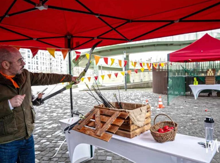 Weihnachtsfeier Outdoor-Winter-Team-Olympiade-Winterliche-Teambuilding-Event Ein Teilnehmer zielt mit Pfeil und Bogen auf einem überdachten Outdoor-Spielfeld einer Winter-Team-Olympiade. Unter roten Pavillons stehen Holzkisten mit Pfeilen, bunte Wimpelketten schmücken den Bereich, und auf einem Tisch liegen Deko-Äpfel in einem Korb. Die Szene zeigt eine winterliche, spielerische Station für eine Outdoor-Weihnachtsfeier.