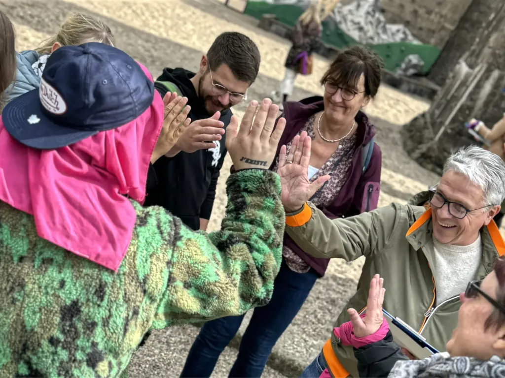 Mehrere Teilnehmende eines Teamevents geben sich in einer lockeren Outdoor-Szene ein gemeinsames High-Five. Die Gruppe steht fröhlich beisammen, einige tragen farbige Tücher oder Jacken, und im Hintergrund ist eine sandige Spielfläche mit Kletterwand zu sehen. Die Stimmung wirkt motiviert, aktiv und teamorientiert.