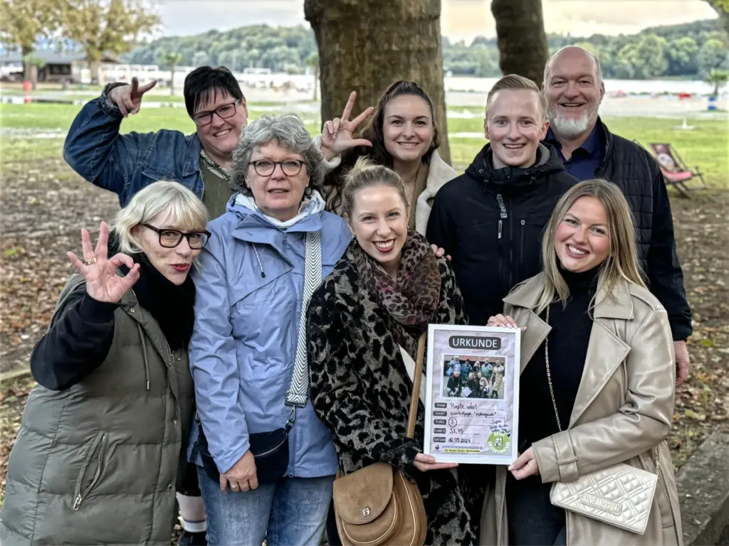 Eine Gruppe fröhlicher Teilnehmender posiert im Freien für ein Erinnerungsfoto nach einem Teamevent. Die Gruppe hält gemeinsam eine Urkunde und steht vor einer herbstlichen Parkkulisse mit Bäumen und See im Hintergrund. Die Stimmung wirkt ausgelassen und stolz auf das erreichte Ergebnis.