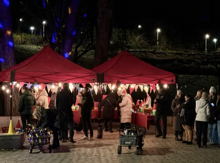 Glühweinwanderung-Firmenfeier-Weihnachtsfeier-Winterevent-Glühweintour Winterliches Glühwein-Event im Freien: Unter zwei rot beleuchteten Pavillons treffen sich Gruppen zu einer Glühweinwanderung, umgeben von Lichterketten, weihnachtlicher Dekoration und festlich geschmückten Bollerwagen.