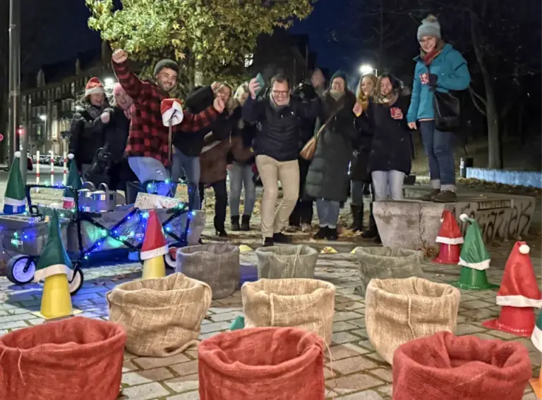 Glühweinwanderung-Firmenfeier-Weihnachtsfeier-Winterliche-Glühweinrallye Winterliches Glühwein-Event mit Spielstation: Eine Gruppe feiert bei einer Glühweinwanderung und steht jubelnd hinter weihnachtlich dekorierten Spielkörben, während ein festlich beleuchteter Bollerwagen daneben steht.