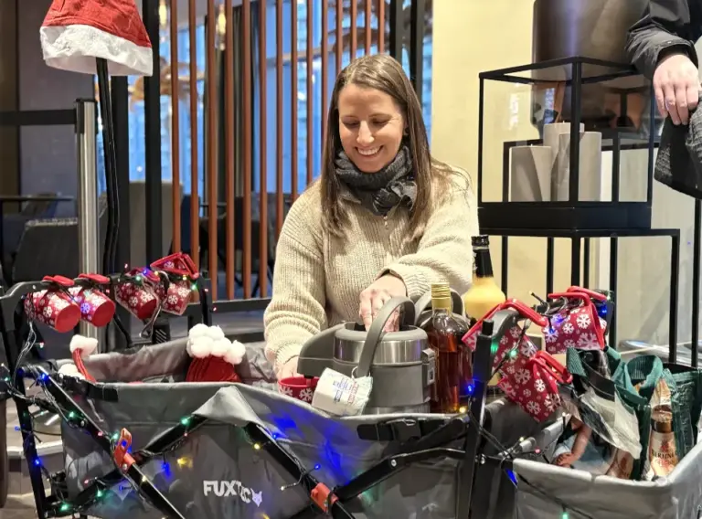 Glühweinwanderung-Weihnachtsfeier-Glühweinrallye Vorbereitung für die Glühweinwanderung: Eine Person schenkt an einem dekorierten Bollerwagen mit Glühweinbehälter, Tassen, Lichterketten und winterlichen Getränken warme Getränke ein.