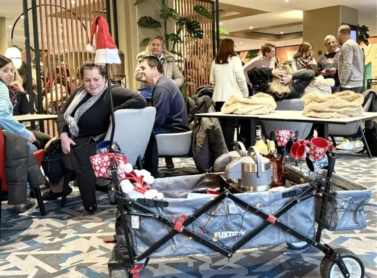 Glühweinwanderung-Weihnachtsfeier-Winterliches-Event-Glühweinrallye Start einer winterlichen Glühweinwanderung: In einem Aufenthaltsbereich sitzen und stehen mehrere Menschen zusammen, während ein weihnachtlich dekorierter Bollerwagen mit Glühwein, Tassen und Getränken für die Glühweinrallye bereitsteht.