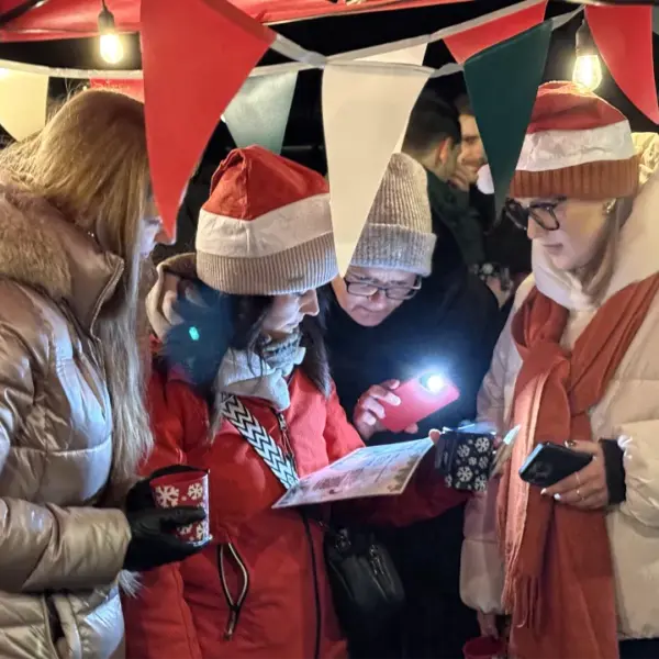 Gruppe bei einer winterlichen Glühweinwanderung: Vier Menschen stehen unter festlicher Beleuchtung und bunten Wimpeln, trinken heißen Glühwein und lesen gemeinsam eine Rallye-Aufgabe während eines winterlichen Team-Events.