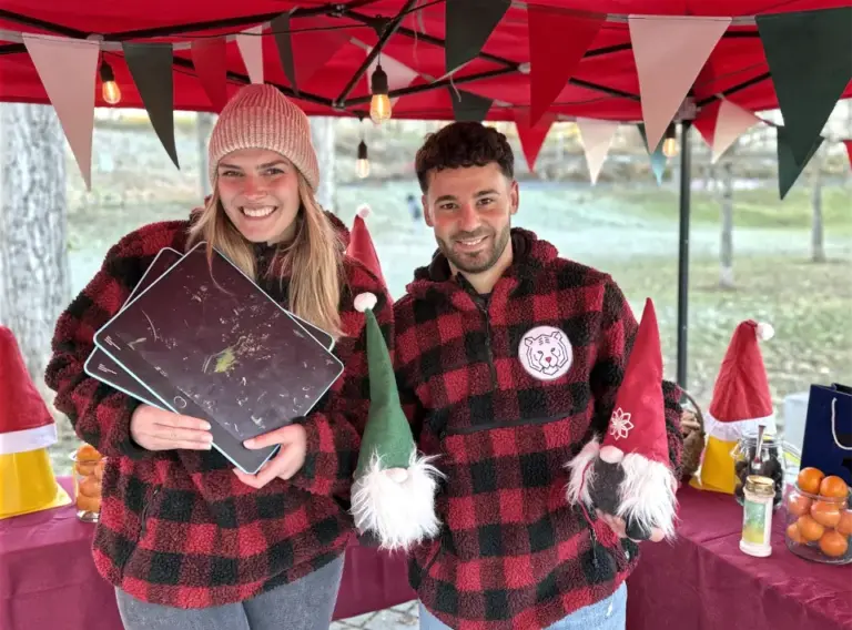 Glühweinwanderung-Weihnachtsfeier-Winterliches-Event-mobile-Spielstation Teammitglieder an einer Spielstation der Glühweinrallye: Zwei Personen in rot-schwarz karierten Winterpullovern stehen unter einem weihnachtlich dekorierten Pavillon und halten Spielmaterialien sowie kleine Wichtelfiguren.