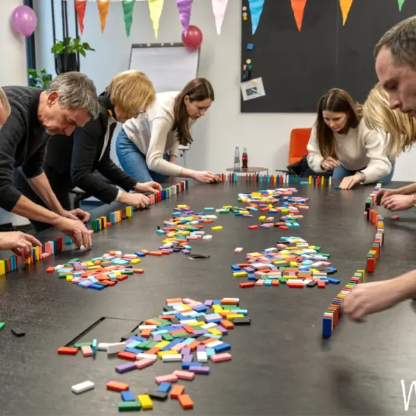 Mehrere Teilnehmende eines Teamevents arbeiten konzentriert an einer langen Dominosteinketten-Challenge. Auf einem großen Tisch werden farbenfrohe Dominosteine gemeinsam aufgestellt, um eine kreative Kettenreaktion zu bauen. Die Aktivität vermittelt Teamwork, Kommunikation und gemeinsames Problemlösen in einer motivierenden und spielerischen Atmosphäre.