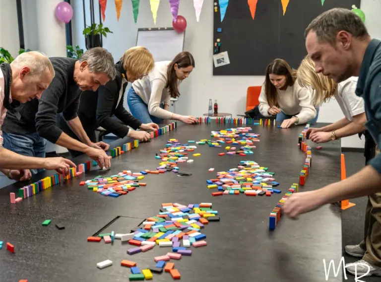 Weihnachtsfeier-Firmenfeier-Teambuilding-Teamevents-Dominoschlange Mehrere Teilnehmende eines Teamevents arbeiten konzentriert an einer langen Dominosteinketten-Challenge. Auf einem großen Tisch werden farbenfrohe Dominosteine gemeinsam aufgestellt, um eine kreative Kettenreaktion zu bauen. Die Aktivität vermittelt Teamwork, Kommunikation und gemeinsames Problemlösen in einer motivierenden und spielerischen Atmosphäre.