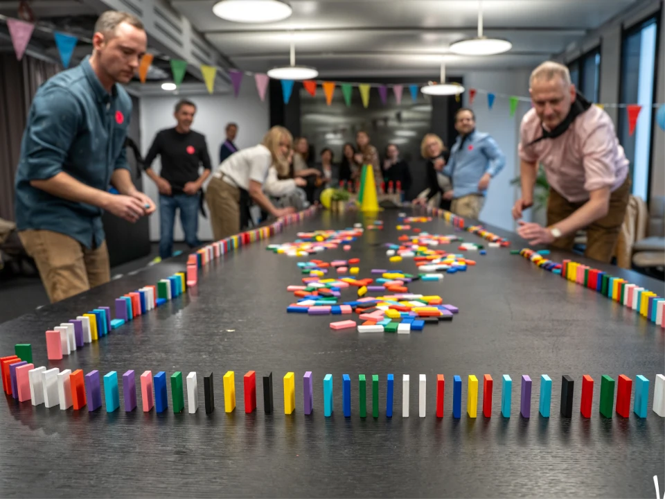Das Foto zeigt ein Team-Building-Spiel im Rahmen einer Indoor-Winter-Olympiade während einer Weihnachtsfeier. Mehrere Personen stehen um einen großen Tisch und bauen gemeinsam eine farbenfrohe Dominostrecke auf. In der Mitte des Tisches liegen zahlreiche bunte Dominosteine, während am Rand bereits eine lange, sorgfältig aufgestellte Kette verläuft. Im Hintergrund ist eine Gruppe weiterer Teilnehmer zu sehen, begleitet von bunter Wimpeldekoration und fröhlicher Stimmung. Die Szene vermittelt Teamarbeit, Konzentration und spielerischen Wettkampf – ein dynamischer Programmpunkt der festlichen Veranstaltung.