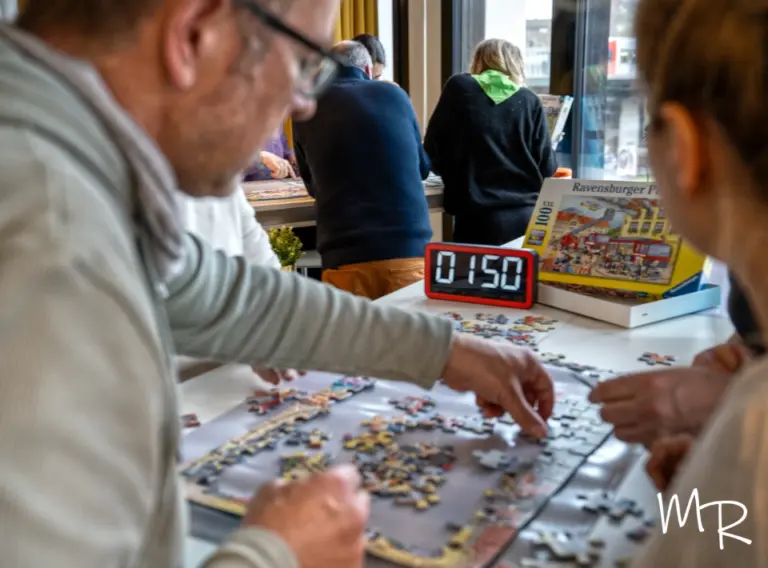 Weihnachtsfeier-Winterevents-Teambuilding-Puzzlechamp Teilnehmende einer winterlichen Firmenfeier arbeiten gemeinsam an einer Puzzle-Challenge, bei der ein Timer den weihnachtlichen Teamwettbewerb begleitet. Auf dem Tisch wird konzentriert ein großes Ravensburger Puzzle zusammengesetzt, während weitere Gruppen im Hintergrund an eigenen Aufgaben arbeiten. Die Szene zeigt gemeinsames Rätseln, Kommunikation und Teamgeist in einer gemütlichen, winterlichen Event-Atmosphäre – ideal für Weihnachtsfeiern und Indoor-Teambuilding im Winter.