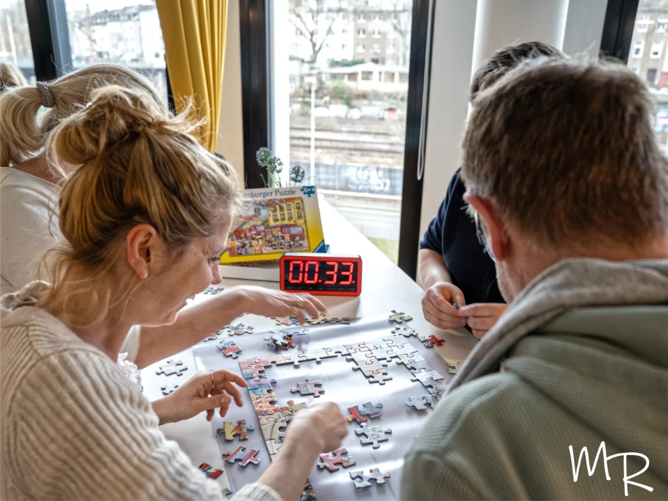 Das Foto zeigt ein Team, das im Rahmen einer weihnachtlichen Indoor-Winter-Olympiade gemeinsam an einem Puzzle arbeitet. Mehrere Personen sitzen konzentriert um einen Tisch und fügen Puzzleteile zusammen, während eine digitale Uhr die verbleibende Zeit für die Challenge anzeigt. Durch die großen Fenster fällt Tageslicht in den festlich geschmückten Veranstaltungsraum der Weihnachtsfeier. Die Szene vermittelt Zusammenarbeit, Spannung und fröhliche Wettkampfstimmung – typisch für ein festliches Team-Building-Event in der Adventszeit.