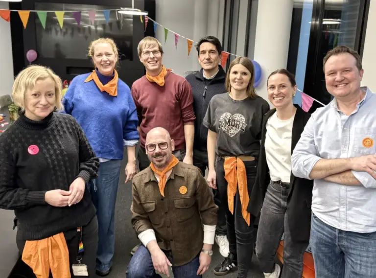 Winterteambuilding-Winterteamolympiade-Weihnachtsfeier-Indoor Das Foto zeigt eine fröhliche Gruppe von Teilnehmenden einer weihnachtlichen Indoor-Winter-Olympiade. Die Personen stehen gemeinsam vor bunten Wimpelketten und tragen orangefarbene Teamtücher oder Teammarkierungen, was auf eine abgeschlossene Challenge oder ein Teamfoto nach dem Event hindeutet. Die Gruppe lächelt in die Kamera und vermittelt Teamgeist, gute Stimmung und ein positives Miteinander. Im Hintergrund sind Teile des festlich dekorierten Veranstaltungsraums zu sehen.