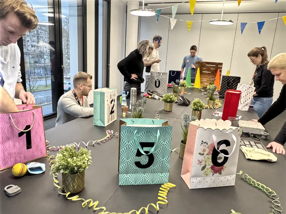 Das Foto zeigt eine Gruppe von Teilnehmern, die im Rahmen einer weihnachtlichen Indoor-Winter-Olympiade an einer Team-Challenge arbeiten. Auf einem großen Tisch stehen mehrere dekorative Geschenk- und Aufgabentüten, jeweils mit Zahlen markiert. Rundherum liegen verschiedene Materialien und Utensilien, die für die Aufgaben benötigt werden. Zwischen den Tüten sind kleine Pflanzendekorationen und spiralförmige Bänder verteilt, während bunte Wimpelketten im Hintergrund eine festliche Atmosphäre schaffen. Die Teilnehmenden sind konzentriert und aktiv in ihre Aufgaben vertieft – eine Szene, die Zusammenarbeit, Kreativität und spielerischen Wettkampf während einer Weihnachtsfeier vermittelt.