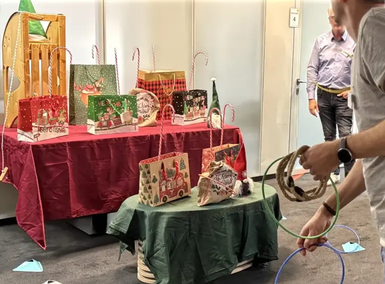 Das Foto zeigt eine Spielstation der weihnachtlichen Indoor-Winter-Olympiade, bei der die Teilnehmenden Ringe auf Zuckerstangen werfen. Auf zwei weihnachtlich dekorierten Tischen stehen Geschenk- und Aufgabentüten, kleine Wichtelfiguren und mehrere aufrecht stehende Zuckerstangen, die als Ziele dienen. Ein Teilnehmer im Vordergrund hält gerade bunte Wurfringe bereit, während weitere Personen im Hintergrund die Szene beobachten. Die festliche Dekoration aus roten und grünen Stoffen sowie die Weihnachtsmotive auf den Geschenktüten schaffen eine warme, spielerische Atmosphäre.