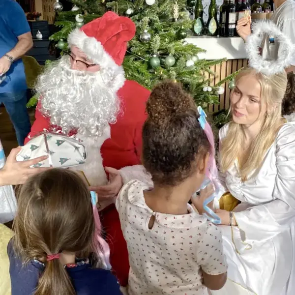 Weihnachtsmann buchen oder Weihnachtsengel mieten bei dem Kinder dem Weihnachtsmann ein Geschenk überreichen, während ein Weihnachtsengel freundlich zusieht – festlicher Besuch beim Kindergeburtstag.