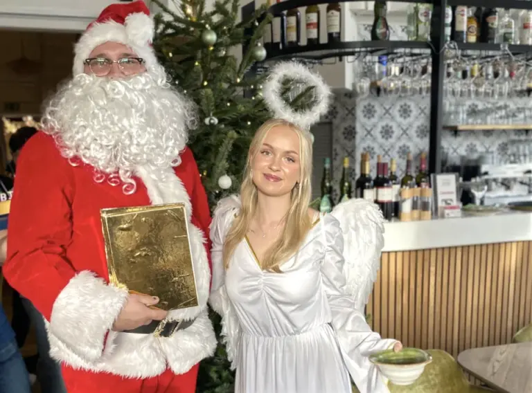 Weihnachtsmann mieten und Weihnachtsengel mieten bei einer Weihnachtsfeier – festlicher Duo-Auftritt Weihnachtsmann mieten und Weihnachtsengel stehen festlich gekleidet vor einem geschmückten Weihnachtsbaum während einer Weihnachtsfeier.