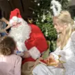 Weihnachtsmann und Weihnachtsengel sitzen vor einem geschmückten Weihnachtsbaum und sprechen mit Kindern, während der Weihnachtsmann ein Geschenk überreicht.