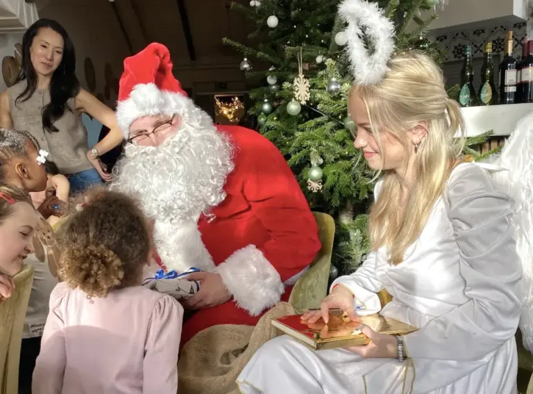 weihnachtsmann-engel-kinder-weihnachtsfeier-geschenk-auftritt.webp Weihnachtsmann und Weihnachtsengel sitzen vor einem geschmückten Weihnachtsbaum und sprechen mit Kindern, während der Weihnachtsmann ein Geschenk überreicht.