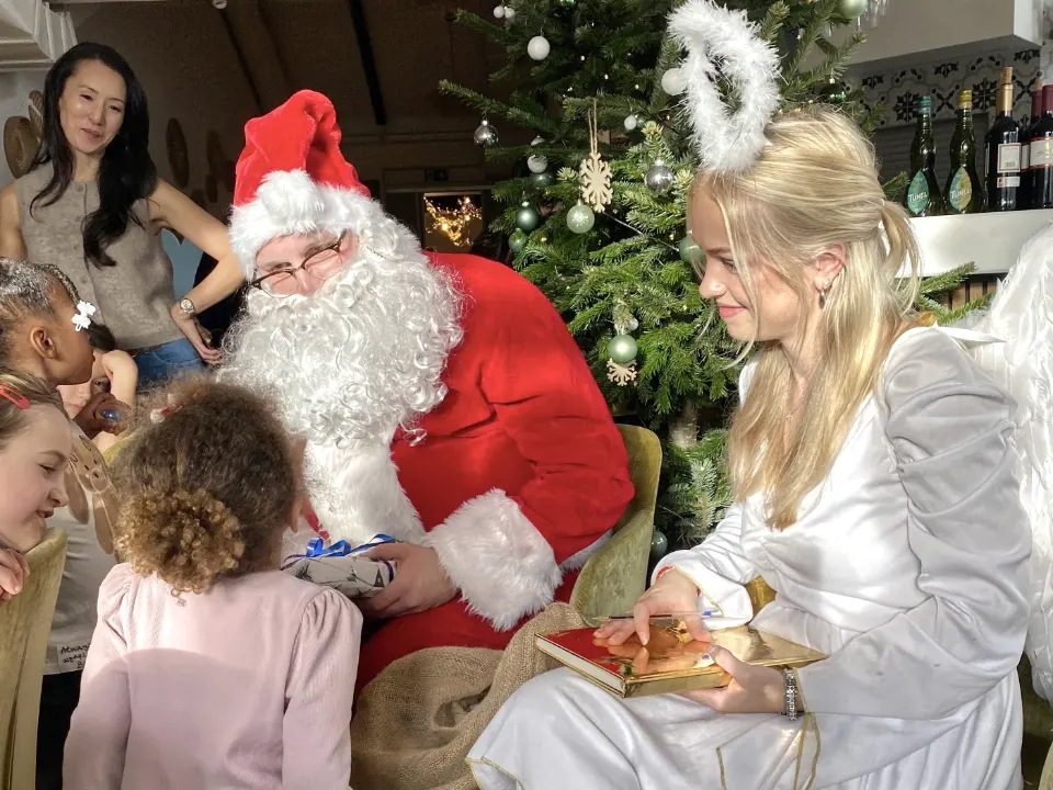 Weihnachtsmann und Weihnachtsengel sitzen vor einem geschmückten Weihnachtsbaum und sprechen mit Kindern, während der Weihnachtsmann ein Geschenk überreicht.