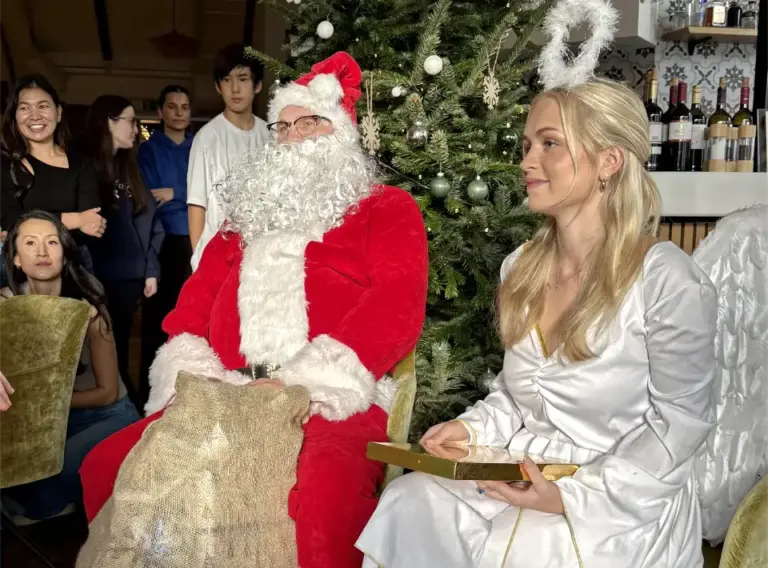 Weihnachtsmann und Weihnachtsengel bei einer Weihnachtsfeier – festlicher Auftritt mit Publikum Weihnachtsmann und Weihnachtsengel sitzen vor einem geschmückten Weihnachtsbaum, umgeben von Gästen während einer Weihnachtsfeier.