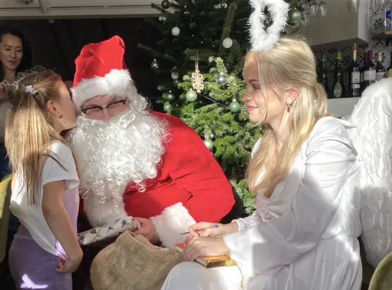 weihnachtsmann-engel-kind-weihnachtsfeier-fluestern-geschenk.webp Kind flüstert dem Weihnachtsmann bei einer festlichen Weihnachtsfeier etwas zu, während ein Weihnachtsengel daneben sitzt und die Szene begleitet.