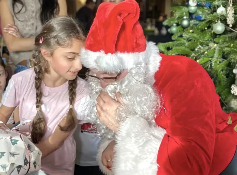 weihnachtsmann-kind-geschenk-weihnachtsfeier-interaktion-walking-act Weihnachtsmann hört einem Kind bei einer festlichen Weihnachtsfeier zu, während das Mädchen ihm etwas ins Ohr flüstert und ein Geschenk hält.