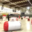 Mitarbeitende spielen mobiles Arrow Tag bei einem Indoor-Firmenevent in einer Sporthalle zwischen aufblasbaren Hindernissen