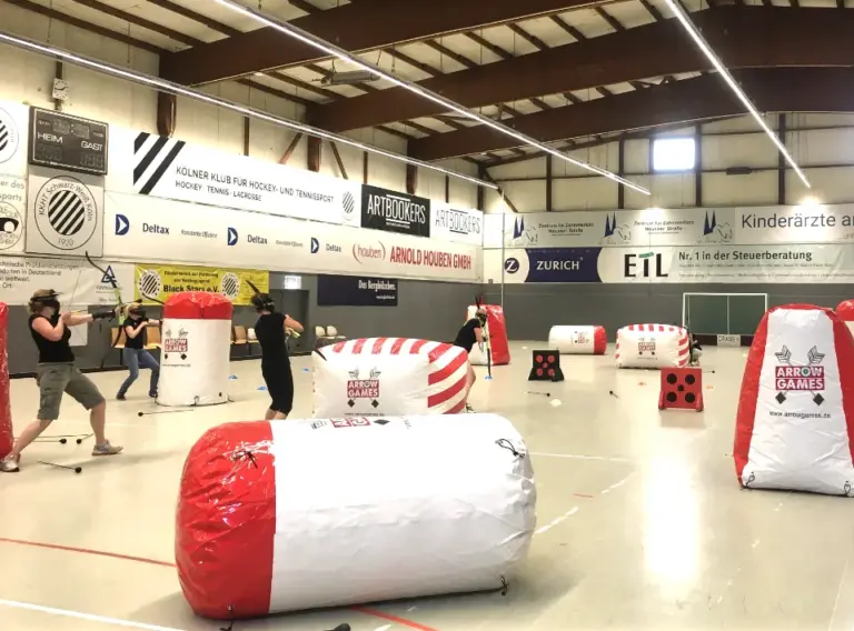 Indoor Arrow Tag für Unternehmen – Actionreiches Firmenevent in der Halle Mitarbeitende spielen mobiles Arrow Tag bei einem Indoor-Firmenevent in einer Sporthalle zwischen aufblasbaren Hindernissen